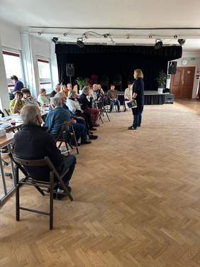 Pokaż zdjęcie: W sali na krzesłach siedzą starsi ludzie. Przed nimi stoi kobieta, która do nich coś mówi. W dłoni trzyma poradniki. 