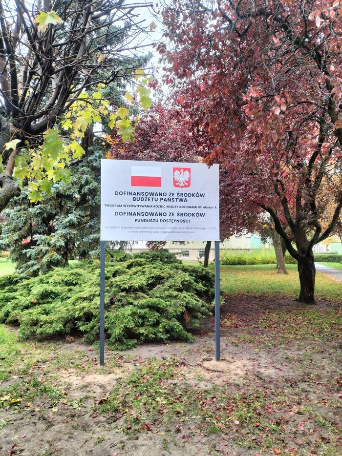 Pokaż zdjęcie: Tablica informacyjna ustawiona na terenie zadrzewionego skweru. Na tablicy znajdują się flagi Polski i godło państwowe oraz informacja o dofinansowaniu inwestycji ze środków budżetu państwa i funduszu dostępności w ramach programu Wyrównywania różnic między regionami. W tle widać drzewa w jesiennych barwach.