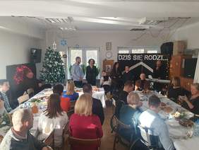 Pokaż zdjęcie: Za stołami siedzą goście, sala udekorowana świątecznie, w lewym rogu stoi choinka, na scenie trwa występ mieszkańców.