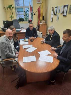 Pokaż zdjęcie: Przy stole siedzi pięć osób: jedna kobieta i czterech mężczyzn. Uśmiechają się. Na stole leżą różne dokumeny, telefon, długopisy, pieczątki. W tle widać flagę Polski i flagę Łodzi. 