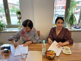 Pokaż zdjęcie: Dwie kobiety w ciemnych włosach i wzorzystych bluzkach siedzą przy drewnianym biurku. Jedna z nich składa podpis na umowę, druga trzyma umowę w ręku. Na biurku leżą pieczątki, kawa i cukierki.