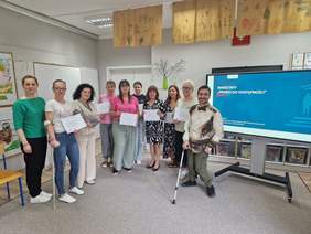 Pokaż zdjęcie: Grupa uczestników warsztatów „Prawo do dostępności” pozuje do wspólnego zdjęcia z certyfikatami, stojąc w sali z ekranem prezentacyjnym i pracami plastycznymi na ścianach.