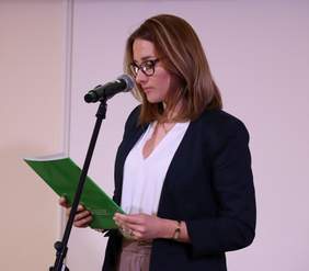 Pokaż zdjęcie: Młoda kobieta w okularach ubrana w białą bluzkę i granatową marynarkę stoi przed mikrofonem. W dłoniach trzyma teczkę z logotypem Państwowego Funduszu Rehabilitacji Osób Niepełnosprawnych wraz z jej zawartością, czyli listem gratulacyjnym.