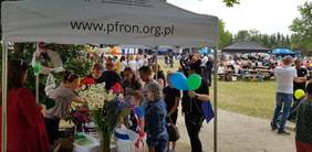 Pokaż zdjęcie: Dwie kobiety stoją pod namiotem z logo PFRON i czytają pytania z zagadnień poruszających tematykę niepełnosprawności dzieciom, które uczestniczą w pikniku. Na stole przykrytym szarym płótnem rozłożone zostały ulotki oraz materiały informacyjne. Dodatkowo na stole ustawione zostały cztery wazony z bukietami polnych kwiatów oraz kosze wiklinowe z ulotkami i gadżetami z logo PFRON. W tle widoczna jest duża scena oraz osoby przemieszczające się pomiędzy stoiskami.
