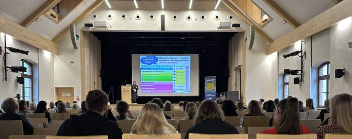 Pokaż zdjęcie: W rzędach krzesła z jasnego drewna na których siedzą ludzie. W tle telebim z prezentacją wysoki skośny jasny sufit. Z nowoczesnym oświtleniem.