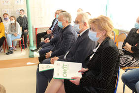 Pokaż zdjęcie: Na krzesłach siedzą osoby wsłuchujące się w wystąpienie, na pierwszym planie kobieta w maseczce oraz mężczyzna w maseczce i okularach, 