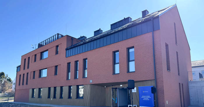 Pokaż zdjęcie: Nowoczesny budynek z czerwonej cegły z dużymi oknami, trzema kondygnacjami i wejściem głównym przygotowanym na wydarzenie – widoczne głośniki i baner informacyjny.