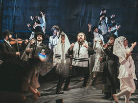 Pokaż zdjęcie: Przedstawienie teatralne. Na scenie grupa mężczyzn i kobiet w strojach żydowskich gra, śpiewa i tańczy.