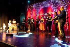 Pokaż zdjęcie: Zdjęcie grupowe wykonane podczas Gali Tęczowych Serc. Uczestnicy stoją na tle ściany wypełnionej zdjęciami dokumentującymi różne wydarzenia i aktywności. Na zdjęciu obecni są m.in. organizatorzy i wręczający nagrody, w tym Dyrektor Oddziału Zachodniopomorskiego PFRON. Jedna z kobiet w różowo-czarnej sukni siedzi z przodu.