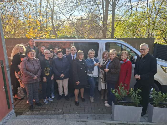 Pokaż zdjęcie: Uczestnicy i kadra WTZ oraz urzędnicy i Dyrektor Oddziału Lubuskiego PFRON stoją na tle auta, a Dyrektor Oddziału przekazuje kluczyki do samochodu