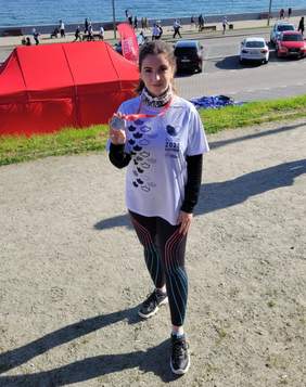 Pokaż zdjęcie: Uczestniczka biegu stoi nad brzegiem morza w Gdyni, trzymając medal „Po Prostu Bieg 2025”. Ma na sobie białą koszulkę z logo wydarzenia, legginsy sportowe i buty do biegania. W tle widać czerwony namiot, samochody, spacerowiczów oraz żaglówki na wodzie.