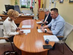 Pokaż zdjęcie: Przy owalnym stole siedzą trzy osoby: kobieta i dwóch mężczyzn. Podpisują umowę. Na stole leżą różne dokumenty, pieczątki, długopisy, telefony.