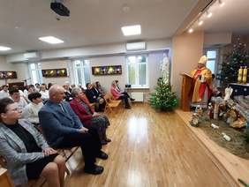 Pokaż zdjęcie: Zdjęcie sali w której odprawiana jest msza święta. Ksiądz biskup głosi Słowo Boże, uczestnicy spotkania siedzą na krzesełkach. Dominuje świąteczny wystrój  sali.rój