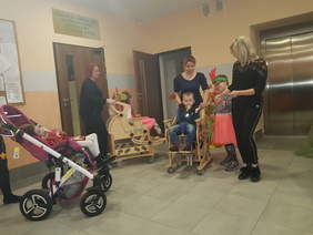 W budynku na tle ściany, drzwi i drzwi windy widzimy czwórkę małych dzieci w tym troje na wózkach w towarzystwie trzech kobiet, zapewne opiekunek