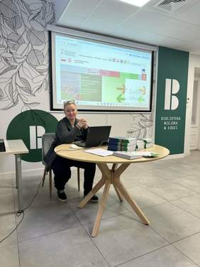 Pokaż zdjęcie: W sali konferencyjnej biblioteki miejskiej trwają warsztaty dotyczące dofinansowań ze środków PFRON. Przy stole siedzi kobieta, która się uśmiecha. W tle znajduje się prezentacja multimedialna. 