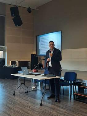 Pokaż zdjęcie: Mężczyzna w garniturze stoi na środku sali. Za nim wyświetlone są informacje na tablicy multimedialnej. 