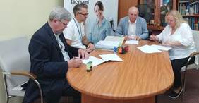 Pokaż zdjęcie: pomieszczenie Oddziału duży stół na którym leżą dokumenty i segregator przy stole siedzi cztery osoby rozmawiają i podpisują dokumenty 