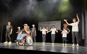 Pokaż zdjęcie: Grupa artystów w czapkach i koszulkach z uśmiechniętymi buźkami występuje na scenie podczas XXVIII Przeglądu Twórczości Artystycznej Osób Niepełnosprawnych w Białymstoku. Na pierwszym planie osoba na wózku śpiewa do mikrofonu, w tle pozostali uczestnicy tańczą.