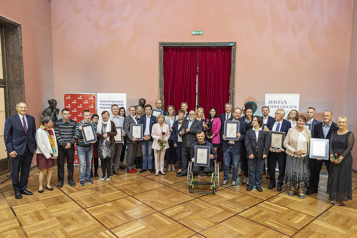 Pokaż zdjęcie: W półokręgu stoją elegancko ubrani,  uśmiechnięci ludzie, część z nich trzyma dyplomy lub kwiaty. Po środku mężczyzna siedzący na wózku inwalidzkim. Stoją na połyskującym parkiecie. Za nimi drzwi z czerwoną kotarą.