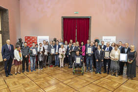 Pokaż zdjęcie: W półokręgu stoją elegancko ubrani,  uśmiechnięci ludzie, część z nich trzyma dyplomy lub kwiaty. Po środku mężczyzna siedzący na wózku inwalidzkim. Stoją na połyskującym parkiecie. Za nimi drzwi z czerwoną kotarą.