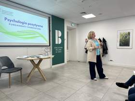 W sali konferencyjnej stoi kobieta. Prowadzi prelekcję. Za nią znajduje się tablica multimedialna, na której jest temat spotkania: "Psychologia pozytywna – fundamenty dobrostanu". 