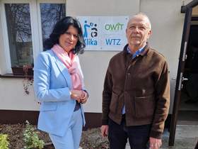 Kobieta i mężczyzna pozują do zdjęcia na tle budynku. W tle na budynku tabliczka z napisami WTZ, Klika, OWiT.