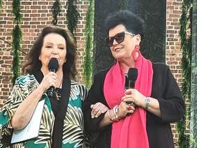 Pokaż zdjęcie: Dwie kobiety: od lewej pani Prezes Anna Dymna oraz pani Bożena Borowiec - Wiceprezes Zarządu PFRON. Kobiety są uśmiechnięte, kolorowo ubrane., trzymają mikrofony, stoją na scenie.