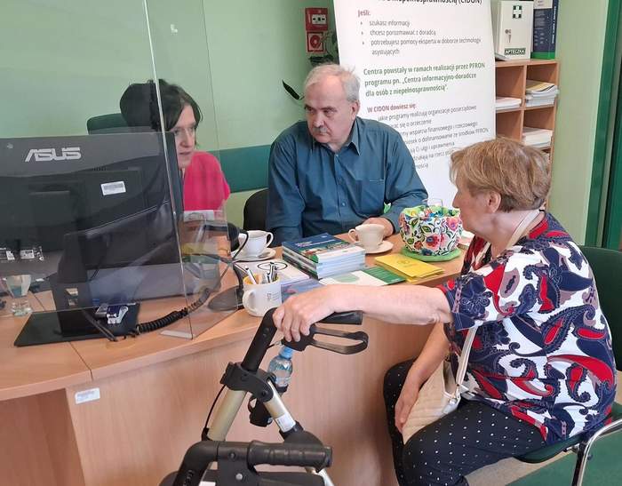 Pokaż zdjęcie: Osoba z chodzikiem rozmawia przy biurku z dwiema osobami w punkcie informacyjnym. Na biurku komputer i ulotki. W tle plakat PFRON dotyczący wsparcia dla osób z niepełnosprawnością.