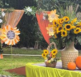 Pokaż zdjęcie: Zdjęcie sceny przyozdobionej kwiatami słoneczników w wazonie oraz dużymi okazami dyni leżącymi obok wazonu.