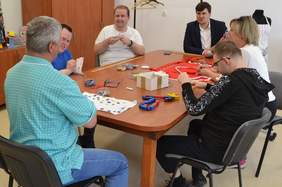 Pokaż zdjęcie: Przy stole siedzi 6 osób. Po ich twarzach widać, że prowadzą rozmowę. Zajmują się krawiectwem