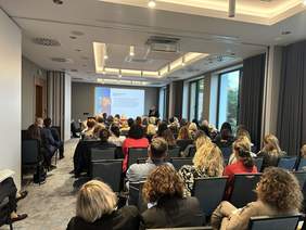 Pokaż zdjęcie: Sala wykładowa, ludzie siedzą rządach na szarych krzesłach przed prowadzącym, za którym znajduje się wielki ekran z prezentacją. Na dole szara wykładzina. Po prawej okna z widokiem na drzewa z długimi zasłonami.