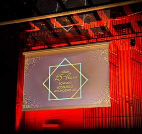Pokaż zdjęcie: Na scenie Filharmonii Łódzkiej zawieszona jest grafika. Na tablicy multimedialnej wyświetlona jest grafika z informacją o 25-leciu Powiatu Łódzkiego Wschodniego. Tło jest w kolorze intensywnie czerwonym. 