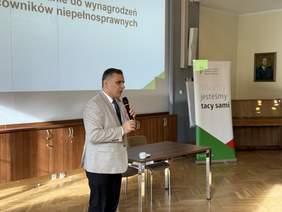 Pokaż zdjęcie: Mężczyzna w jasnej marynarce i ciemnych spodniach przemawia do zgromadzonych, trzymając mikrofon. Za nim widoczny jest baner PFRON z hasłem „Wszyscy jesteśmy tacy sami” oraz slajd prezentacji dotyczącej dofinansowania wynagrodzeń pracowników z niepełnosprawnością.