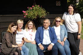 Pokaż zdjęcie: Na zdjęciu na ławce na środku siedzi mężczyzna o ciemnej karnacji ubrany w białą koszulę, granatowe jeansy i niebieską marynarkę. Po jego lewej i prawej stronie siedzi łącznie 5 kobiet. Wszyscy się uśmiechają. Jest słoneczna pogoda. W tle 