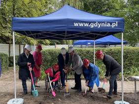 Pokaż zdjęcie: Pod niebieskim, rozkładanym namiotem z napisem Gdynia znajduje się 7 osób w kaskach ochronnych wbijających w ziemie łopaty. Wszyscy są ubrani na galowo. Panuje radosna atmosfera o czym świadczą uśmiechy uczestników.
