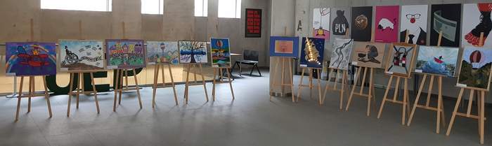 Pokaż zdjęcie: Prace plastyczne oraz plakat promujący wystawę rozmieszczone na trzynastu sztalugach. Wystawa znajduje się w budynku o betonowych murach. Na ścianach zawieszone są różne plakaty. W tle, za wystawą ustawiony jest duży napis: „UEP”.