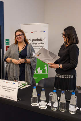 Pokaż zdjęcie: Dwie kobiety od lewej przedstawicielka MZK w Gorzowie Wielkopolskim stoi uśmiechnięta na tle banneru z napisem Różni podróżni – obsługa bez barier. Po prawej p. Dorota Habich Zastępca Prezesa PFRON odczytuje przekazaną, podpisaną deklarację. 