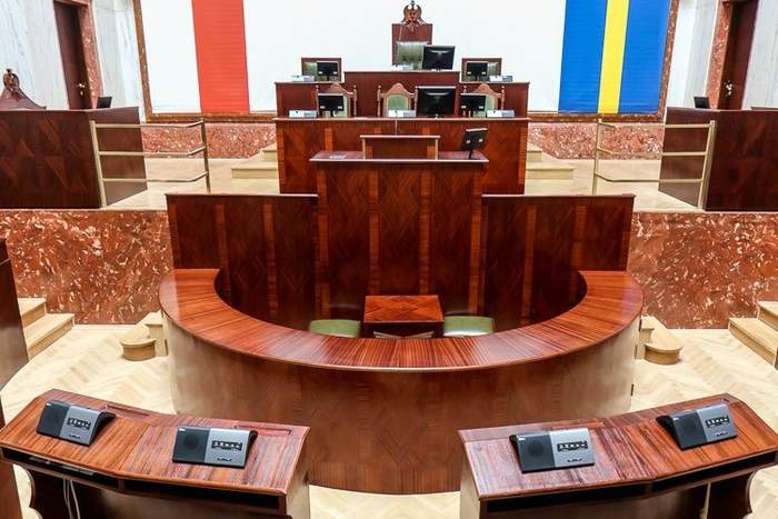 Pokaż zdjęcie: Fragment wnętrza marmurowej sali Sejmu Śląskiego na pierwszym planie drewniane blaty ław na których znajdują się urządzenia do głosowania na środku stoi duża drewniana mównica 
