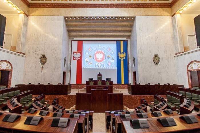 Pokaż zdjęcie: Widok wnętrza bardzo dużej marmurowej sali Sejmu Śląskiego  4 rzędu law ułożone są w półkolu a każdy rząd jest powyżej następnego na blatach znajdują się urządzenia do głosowania na środku stoi duża drewniana mównica 