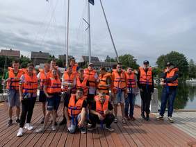 Pokaż zdjęcie: Grupa uczestników warsztatów żeglarskich stojąca na pomoście, ubrana w pomarańczowe kamizelki ratunkowe.