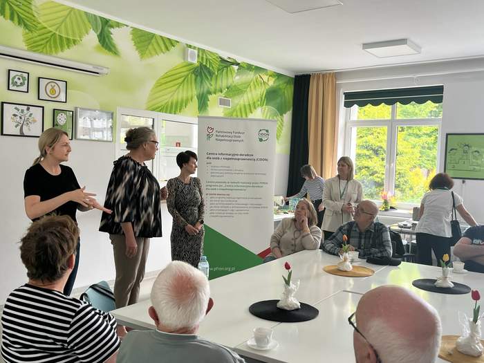 Na środku sali stoja trzy kobiety. Słuchają ich uczestnicy spotkania, siedzący przy stołach. W tle, po prawej stronie widać baner PFRON CIDON