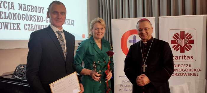 Pokaż zdjęcie: Renata Pieksza wraz z mężem stoją obok biskupa, Renata Pieksza trzyma w ręku nagrodę i różę