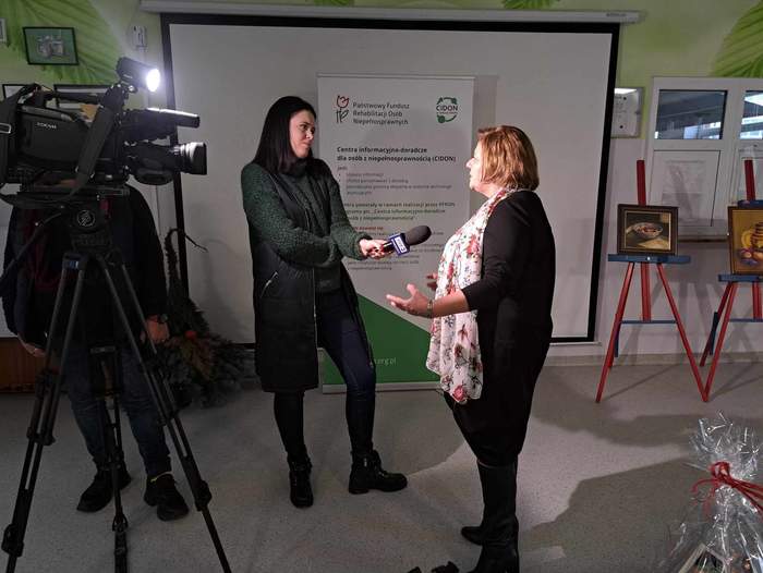 Dyrektor Oddziału PFRON Aleksandra Gierej podczas udzielania wywiadu dla TVP3 Bydgoszcz - od lewej stoi kamera, dziennikarka z mikrofonem w dłoni i  Pani Dyrektor. W tle stoi baner PFRON