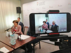 Pokaż zdjęcie: osoba siedząca przy stole odpowiada na pytania trzymając w reku mikrofon, kadr widoczny jest przez ekran smartfona