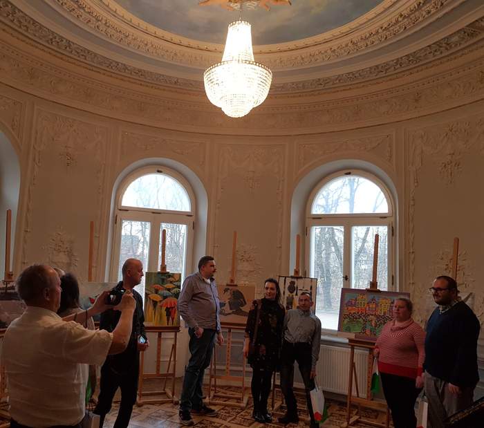 Pokaż zdjęcie: Grupa ośmiu osób stoi w pałacu, w pobliżu wystawy prac plastycznych, z czego część z nich pozuje do zdjęcia na jej tle. W pomieszczeniu, w którym znajduje się wystawa, widoczny jest także podwieszany żyrandol. 