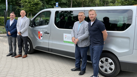 Pokaż zdjęcie: Przy ciemnym mikrobusie z logo PFRON stoi czterech mężczyzn.