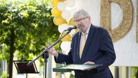 Pokaż zdjęcie: Do mikrofonu wypowiada się dyrektor Łódzkiego Oddziału PFRON. Jest ubrany w garnitur. W dłoni trzyma grawer. W tle widać zieleń. 