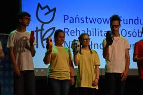 Pokaż zdjęcie: Na scenie w pomieszczeniu stoją dwie dziewczyny i dwóch chłopaków trzymają w ręku instrumenty muzyczne, w tle na dużym ekranie wyświetlone jest logo PFRON