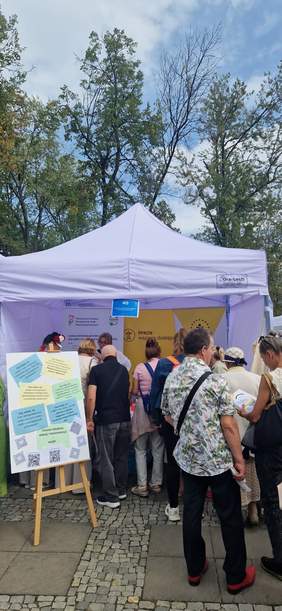 Pokaż zdjęcie: Na zdjęciu widać grupę ludzi stojących w kolejce pod białym namiotem, gdzie odbywa się plenerowe wydarzenie. Przed namiotem po lewej stronie, na sztaludze, umieszczona jest tablica z informacjami w kilku ramkach. W tle widać wysokie drzewa, a nad nimi rozciąga się zachmurzone, błękitne niebo.