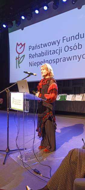 Pokaż zdjęcie: Ubrana na galowo kobieta stoi przy mównicy i przemawia, a za nią logo wyświetlona prezentacja i baner CIDON